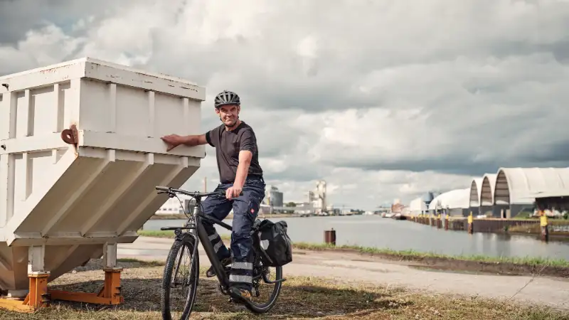 JobRadler in Arbeitshose lehnt an Container, hinter ihm Wasser