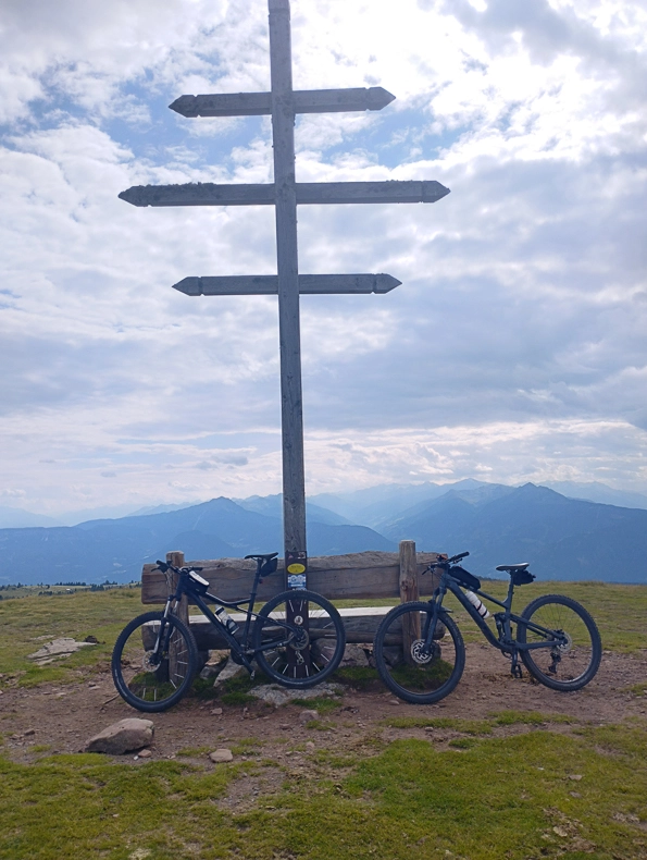 Zwei Mountainbikes stehen vor einer Holzbank unter einem großen Wegweiser auf einem Berggipfel, dahinter wolkiger Himmel und Bergpanorama.