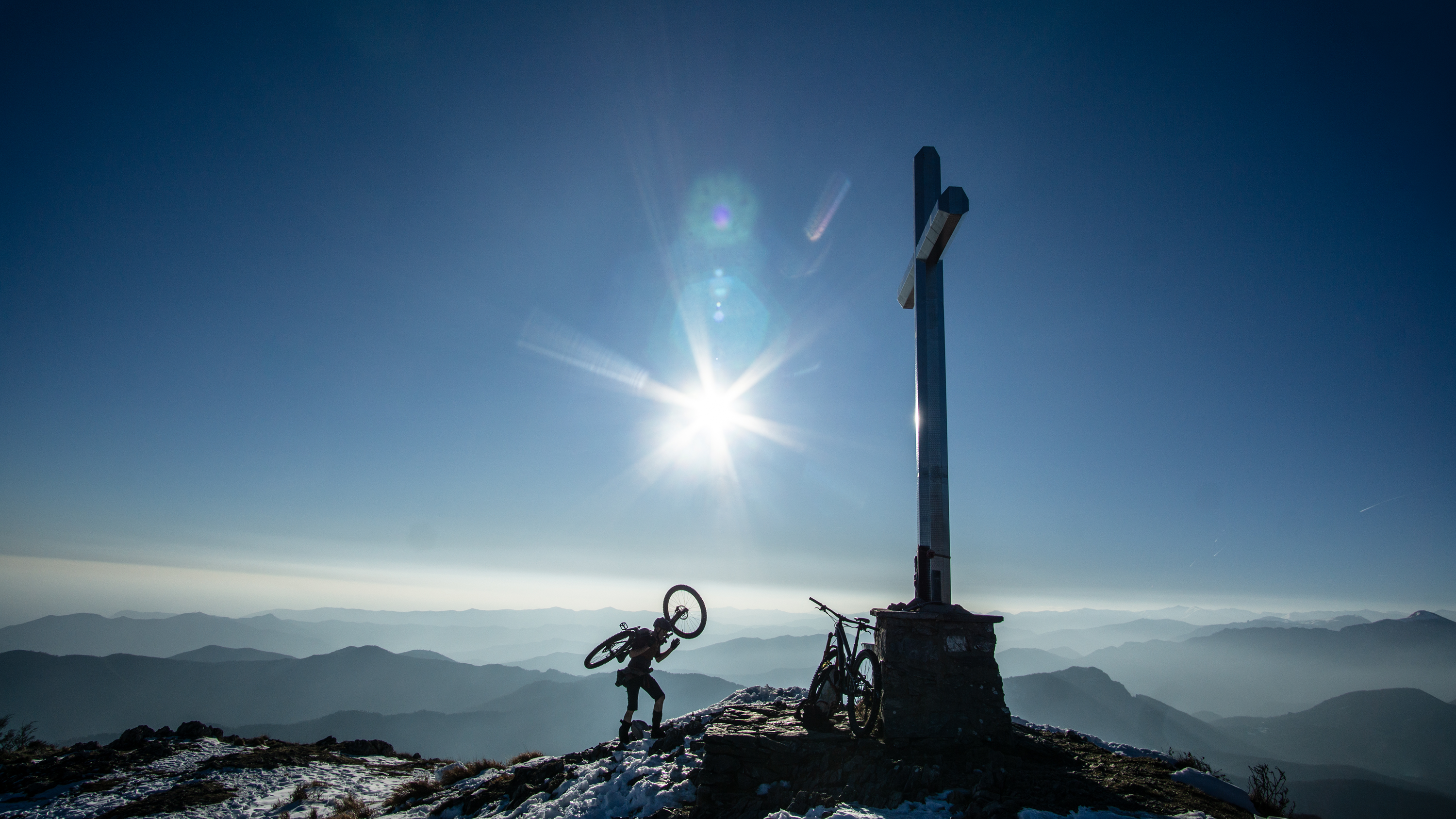 Eine Person steht auf einem verschneiten Berggipfel neben einem großen Kreuz, trägt ein Fahrrad, während ein zweites Rad am Kreuz lehnt; die Sonne beleuchtet die umliegenden Bergketten im Dunst.